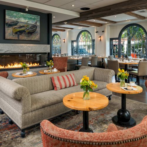 A cozy, elegant restaurant with a fireplace, armchairs, sofas, floral centerpieces, and large windows letting in natural light.