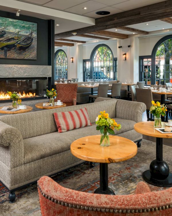 A cozy, elegant restaurant with a fireplace, armchairs, sofas, floral centerpieces, and large windows letting in natural light.