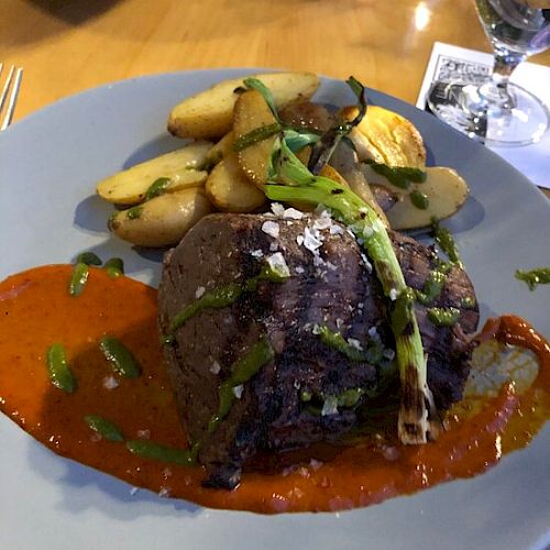 A plate of cooked meat topped with green herbs and grilled vegetables, served with roasted potatoes and a red sauce.