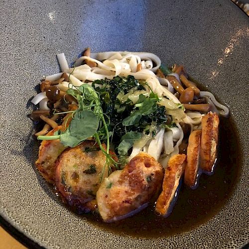 This image shows a bowl of noodle soup topped with greens, mushroom slices, and what appears to be pieces of fried tofu or fish cake.