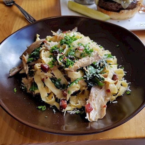 A bowl of pasta topped with greens and pieces of chicken, served on a wooden table with another dish featuring a burger and a pickle in the background.