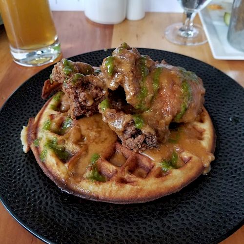 The image shows chicken and waffles on a black plate, accompanied by a glass of beer, a glass of water, and some condiments in the background.