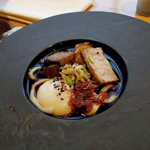 A beautifully plated dish featuring noodles, a soft-boiled egg, slices of meat, vegetables, and garnishes, all in a dark broth.