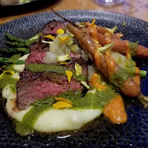 The image shows a plated dish featuring slices of medium-rare steak, baby carrots, mashed potatoes, asparagus, and a green sauce garnished with flower petals.