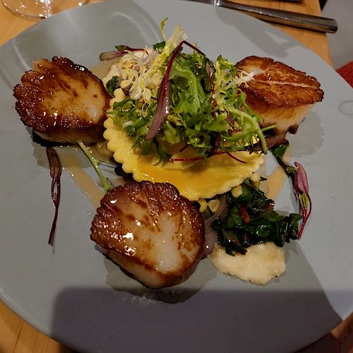 The image shows a plate with seared scallops, a ravioli topped with greens, and a garnish, all served on a light blue plate.