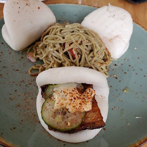 The image shows a plate with three steamed buns, with the bottom one filled, accompanied by a portion of green noodles in the center.