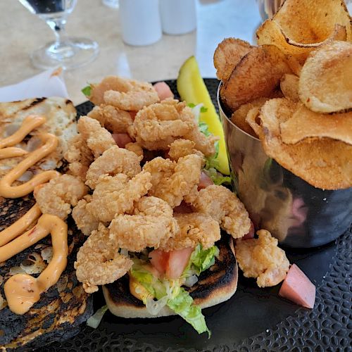 A plate with a shrimp po' boy sandwich topped with lettuce and tomato, a side of crispy potato chips, and a pickle spear.