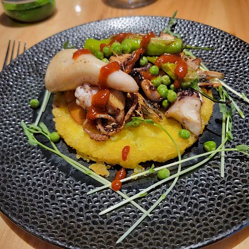 A plated gourmet dish featuring seafood, green peas, sprouts, sauce, and a yellow puree on a textured black plate sits on a wooden table.