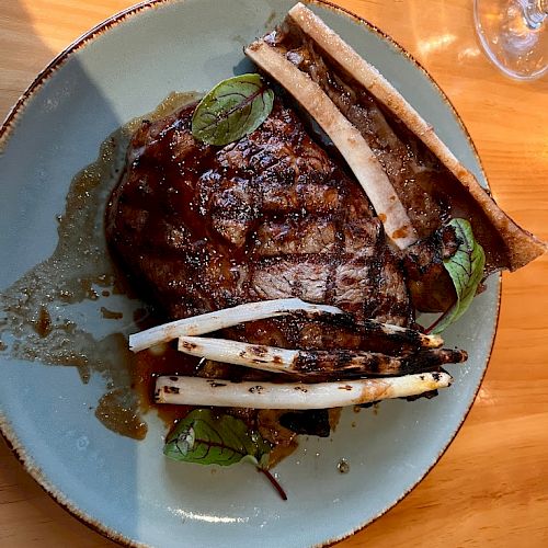 A plate with a grilled steak, garnished with herbs and bones, possibly marrow, on a light blue plate on a wooden table, with a drink nearby.