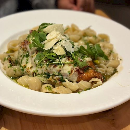 A plate of pasta with a creamy sauce, topped with greens and grated cheese, is on a wooden table, with a person's hand in the background.