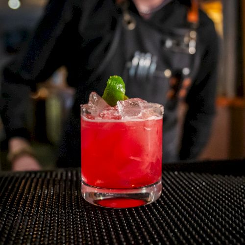 A bartender's hand is seen in the background, focusing on a vibrant red cocktail garnished with a lime wedge on a bar counter with a black mat.