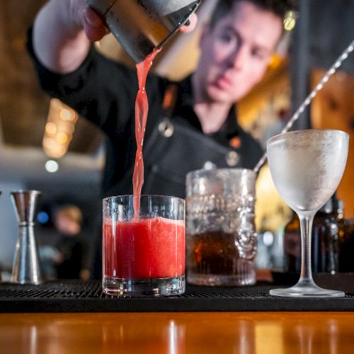 A bartender is pouring a red cocktail into a glass, with various bar tools and a chilled glass next to it.