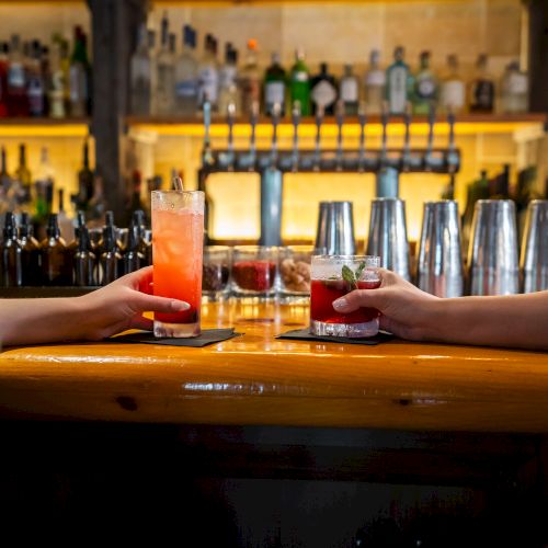 Two people clink glasses with colorful drinks at a bar with various bottles and taps in the background.