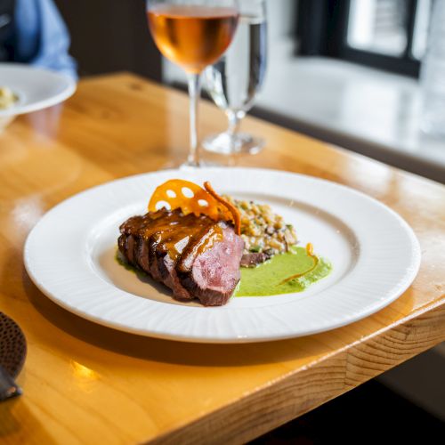 A plated gourmet dish featuring sliced meat, sauce, and garnish is on a wooden table beside a wine glass and water bottle.