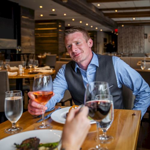 A man in a blue shirt and vest is seated at a restaurant table, holding a glass of rosé wine, smiling at someone across from him.