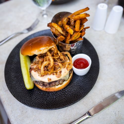 A plate with a burger topped with onion rings, a pickle spear, fries in a metal cup, ketchup, a knife, a fork, and a drink on the side ends the sentence.