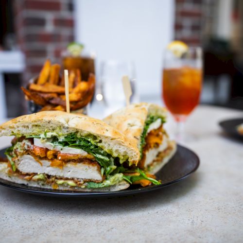 A sandwich with fried chicken, greens, and a side of fries on a table, accompanied by a glass of iced tea.