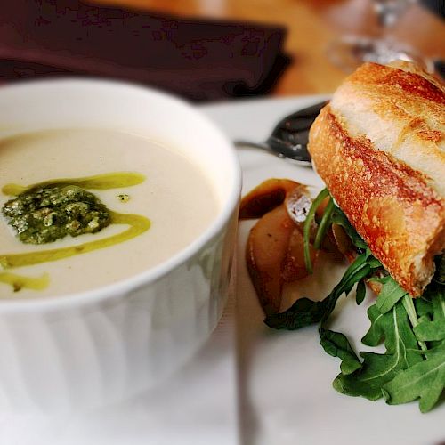 A bowl of creamy soup with pesto garnish alongside a baguette sandwich filled with greens and vegetables, served on a white plate.