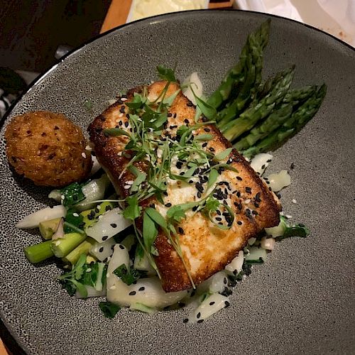 The image shows a plated dish featuring grilled fish topped with herbs, asparagus spears, greens, onions, and a fried ball, likely a croquette.