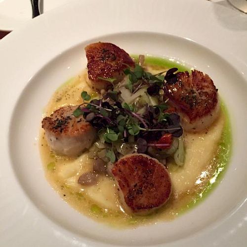 The image shows a gourmet dish of seared scallops on a bed of creamy puree, garnished with microgreens and accompanied by a side of vegetables.