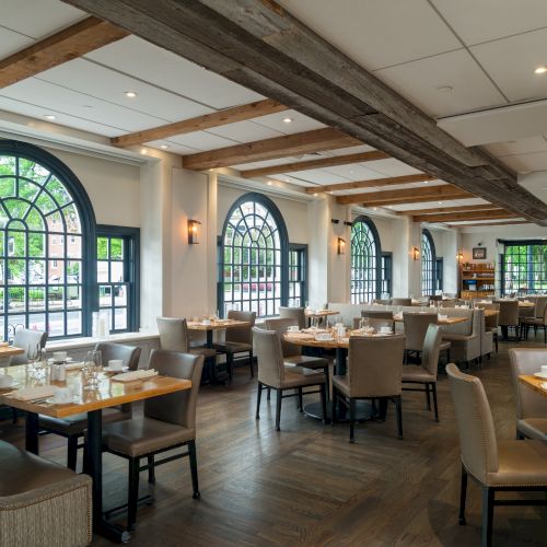 A spacious restaurant with wooden beams, large arched windows, and neatly set tables with gray chairs and white tablecloths, ready for service.