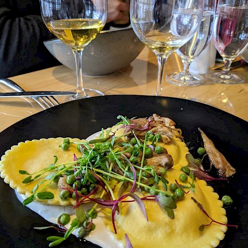 A plate with ravioli topped with microgreens and peas, accompanied by glasses of white wine, is set on a wooden table in a dining setting.