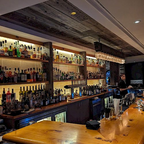 A cozy bar with a wooden counter, shelves stocked with various bottles of alcohol, and a bartender attending to customers in the background.