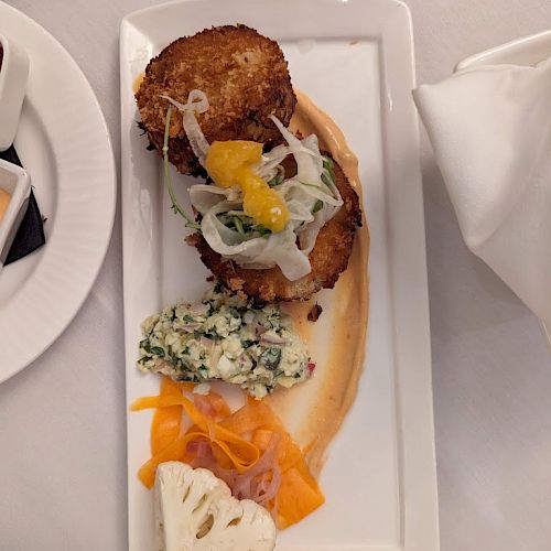 The image shows a plated dish with two pieces of fried food, various vegetables, and a sauce on a rectangular white plate on a white tablecloth.