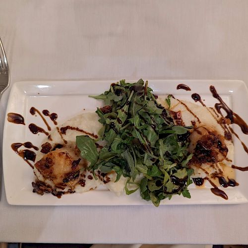 A rectangular white plate with a salad, seared scallops, and a balsamic glaze drizzle, set on a white tablecloth with a fork and knife.