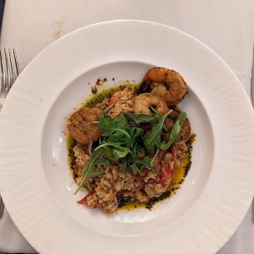 This image shows a plate of food featuring shrimp, grains, and greens, possibly arugula, garnished with herbs and a drizzle of sauce.