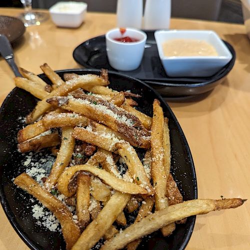 A bowl of seasoned fries with grated cheese accompanied by two dipping sauces, a plate, salt and pepper shakers, and a glass of water on a table.