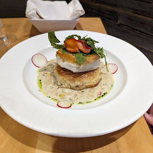 A gourmet dish featuring stacked items on a white plate, topped with greens and accompanied by a creamy sauce, garnished with radish slices.