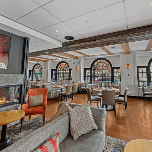 A cozy restaurant interior with a fireplace, modern decor, and large windows, featuring tables and chairs arranged for dining.