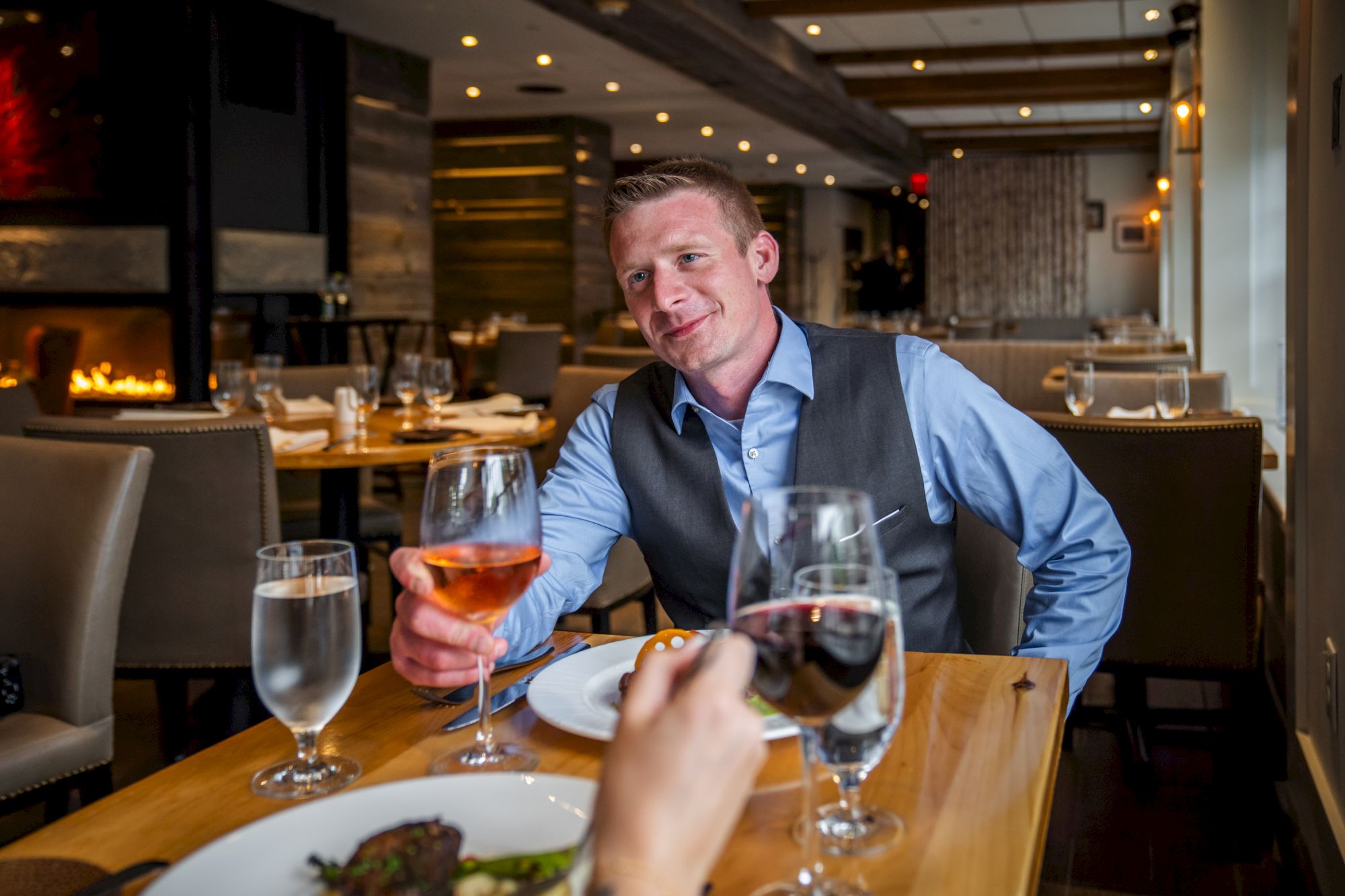 A man in a blue shirt and vest is sitting at a restaurant table, holding a glass of wine, while dining with another person.