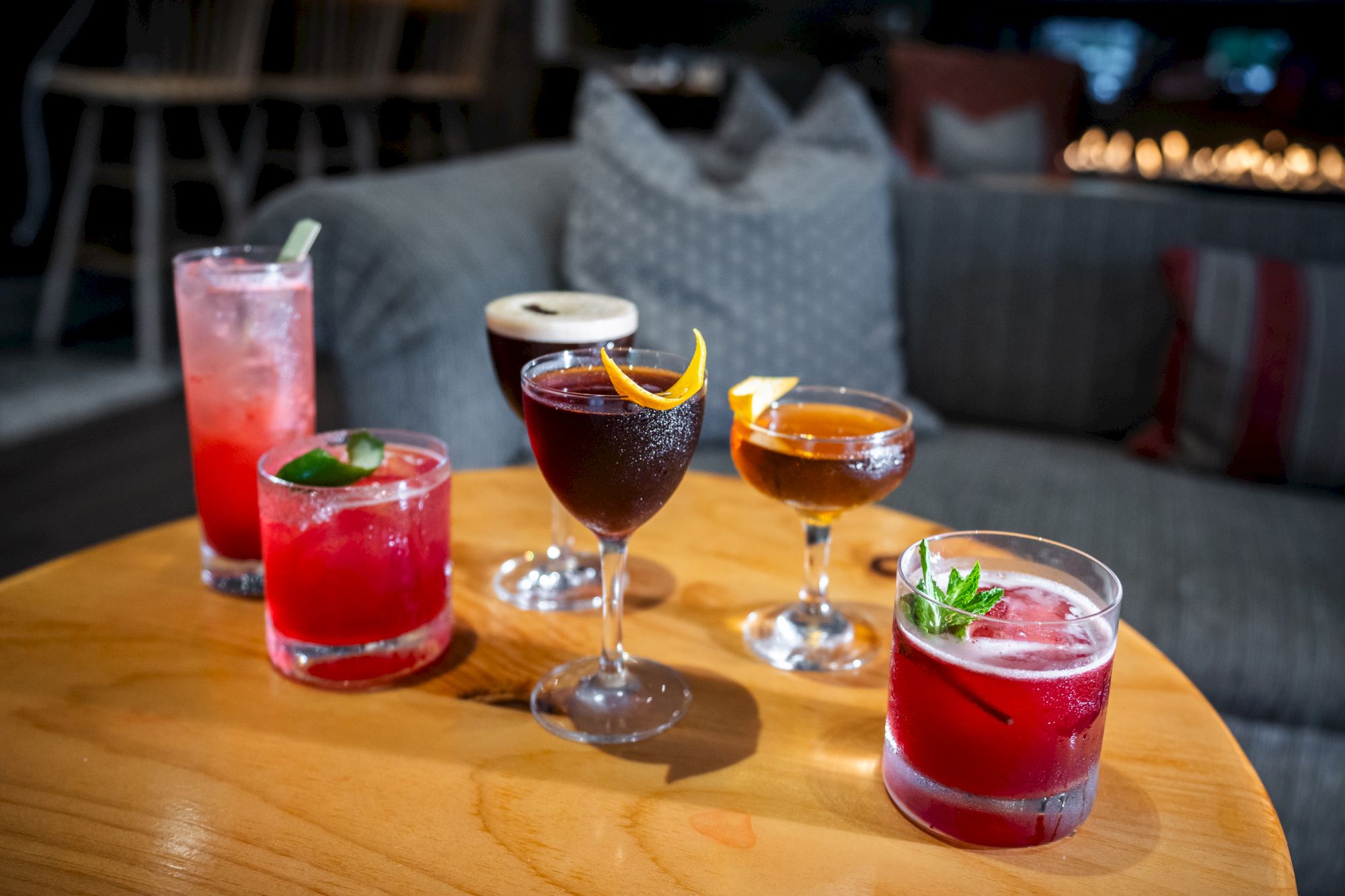 The image shows a variety of colorful cocktails on a wooden table, with a cozy, blurred background that includes cushions and a dimly lit setting.