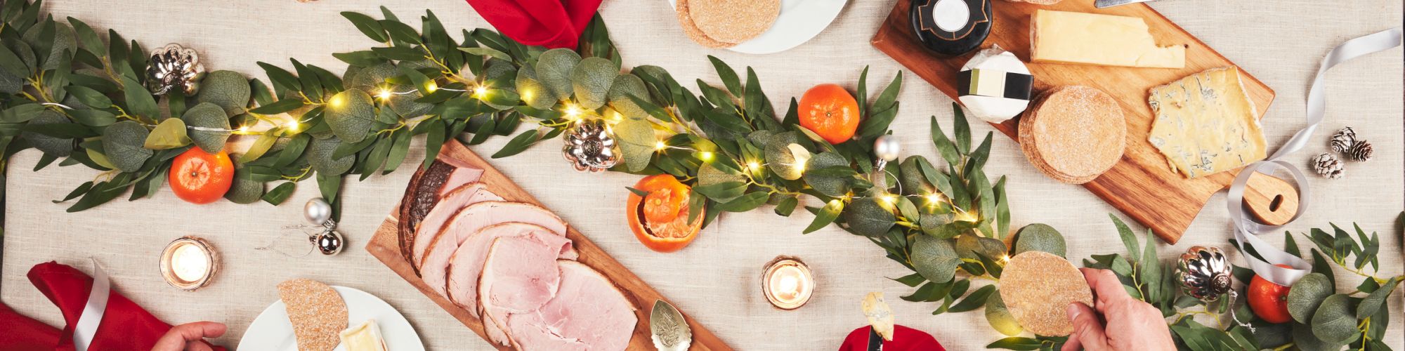 A festive table setting with food, cheese, ham, candles, greenery, and people reaching for various items.