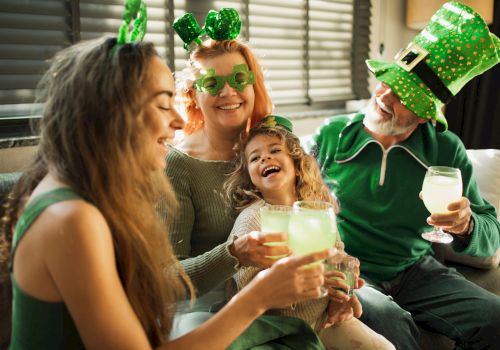 A joyful group celebrates with drinks, wearing green clothes and hats, possibly for St. Patrick's Day, smiling and enjoying together.