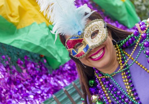 A person wearing a colorful mask and beads, smiling in front of vibrant decorations.