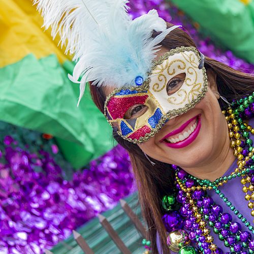 A person wearing a colorful mask and beads, smiling in front of vibrant decorations.