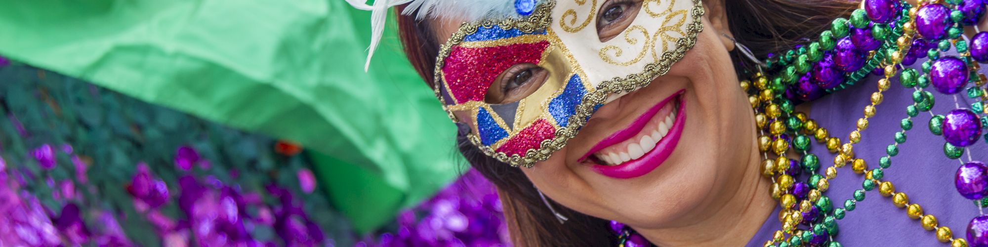 A person is smiling, wearing a colorful feathered mask and Mardi Gras beads. They stand in front of vibrant decorations.