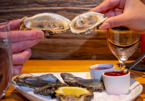 Two hands holding oyster shells above a plate with oysters, lemon, and sauces; glasses of white wine are visible.