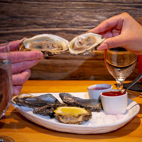 Two hands holding oyster shells above a plate with oysters, lemon, and sauces; glasses of white wine are visible.