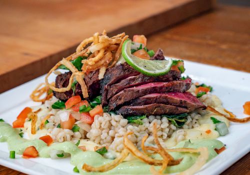 A plated dish with sliced meat, crispy onion strips, vegetables, and a grain base, garnished with sauce, on a wooden table.