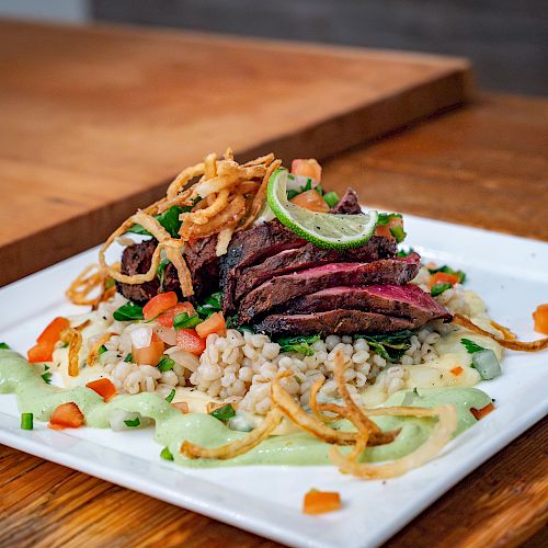 A plated dish with sliced meat, crispy onion strips, vegetables, and a grain base, garnished with sauce, on a wooden table.