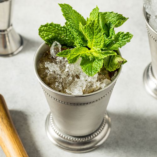 A mint julep cocktail with crushed ice and fresh mint leaves in a metal cup, sitting next to a wooden muddler on a light surface.