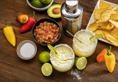 The image shows margaritas with lime, a shaker, salsa, avocados, peppers, and tortilla chips on a wooden table.