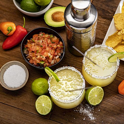 The image shows margaritas with lime, a shaker, salsa, avocados, peppers, and tortilla chips on a wooden table.