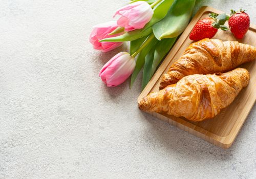 Two croissants on a wooden tray, accompanied by pink tulips and strawberries, placed on a light textured surface.