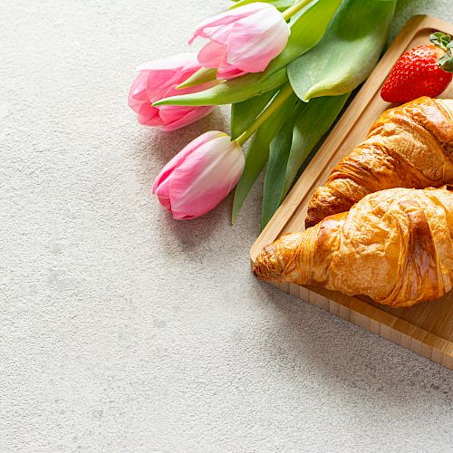 Two croissants on a wooden tray, accompanied by pink tulips and strawberries, placed on a light textured surface.